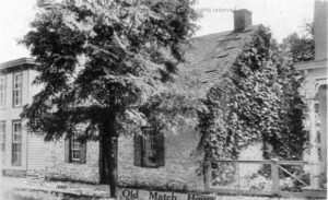 Black and white image of brick building with ivy-covered side and tree in front. Sign with "Old Match House" printed on it is visible